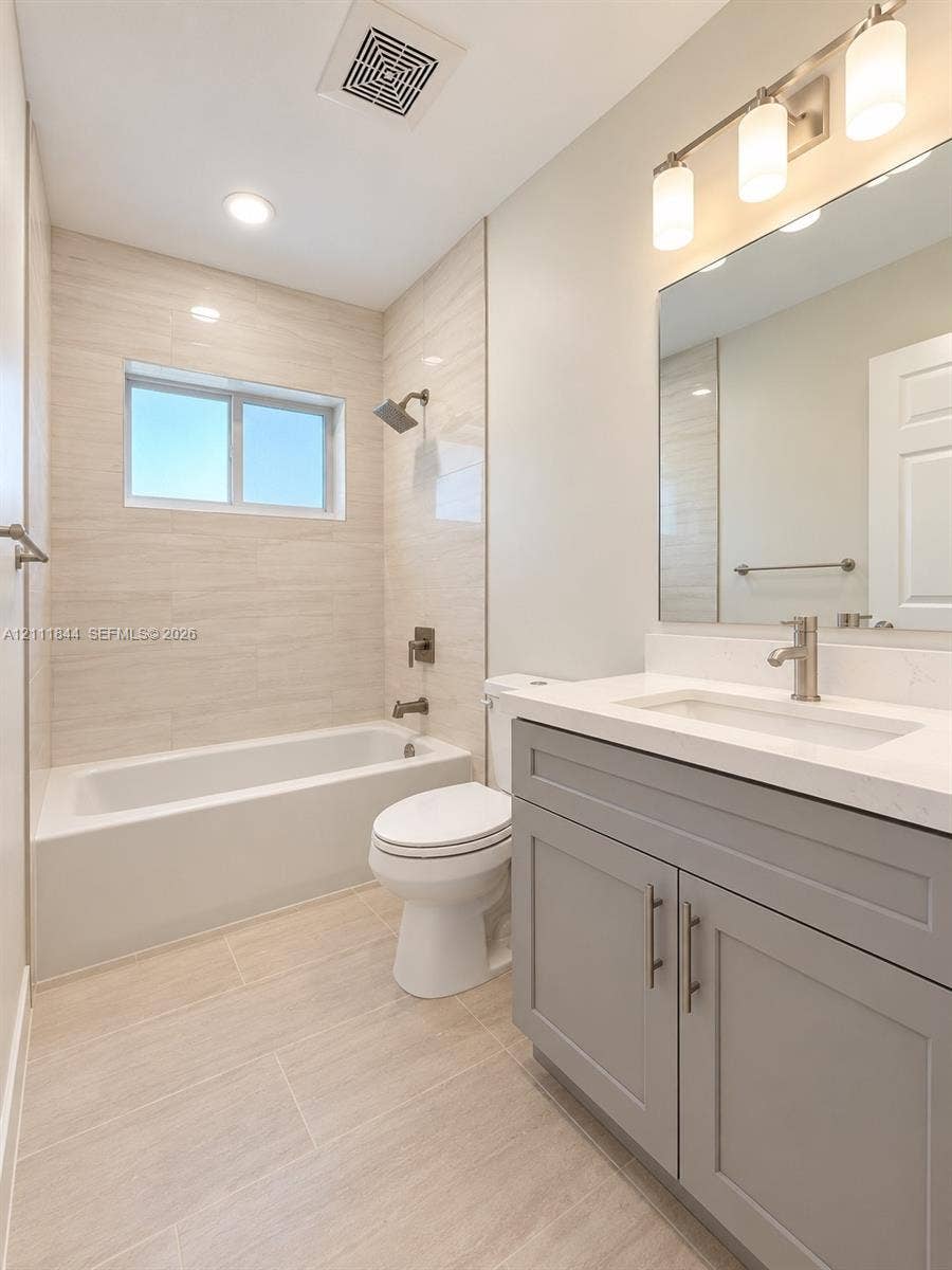 Bathroom featuring a gray vanity with a stone countertop, an integrated rectangular sink, neutral large-format wall and floor tile, and a bathtub with showerhead and mixer faucet