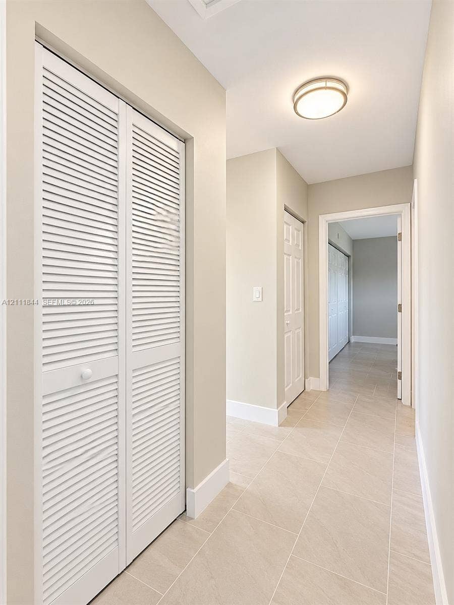 Hallway featuring neutral-toned tile flooring, white louvered-style closet doors, and a flush-mount circular ceiling fixture