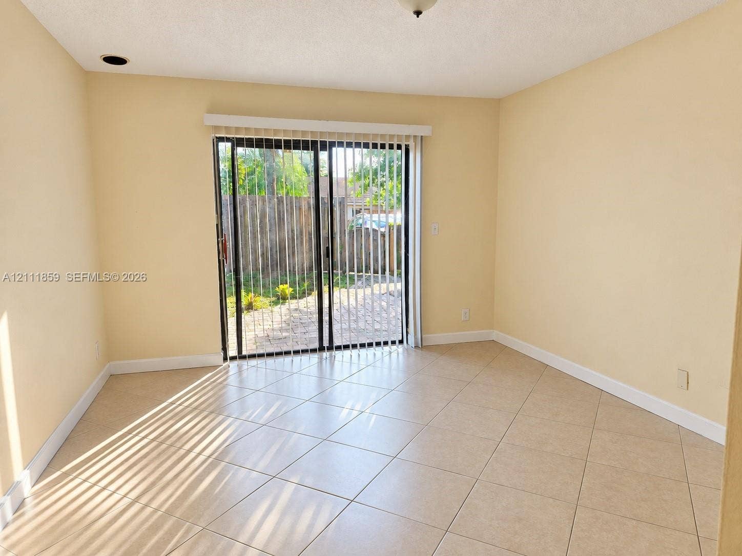 Room featuring ceramic tile flooring, neutral wall paint, and sliding glass doors with vertical blinds providing exterior access to a fenced outdoor area