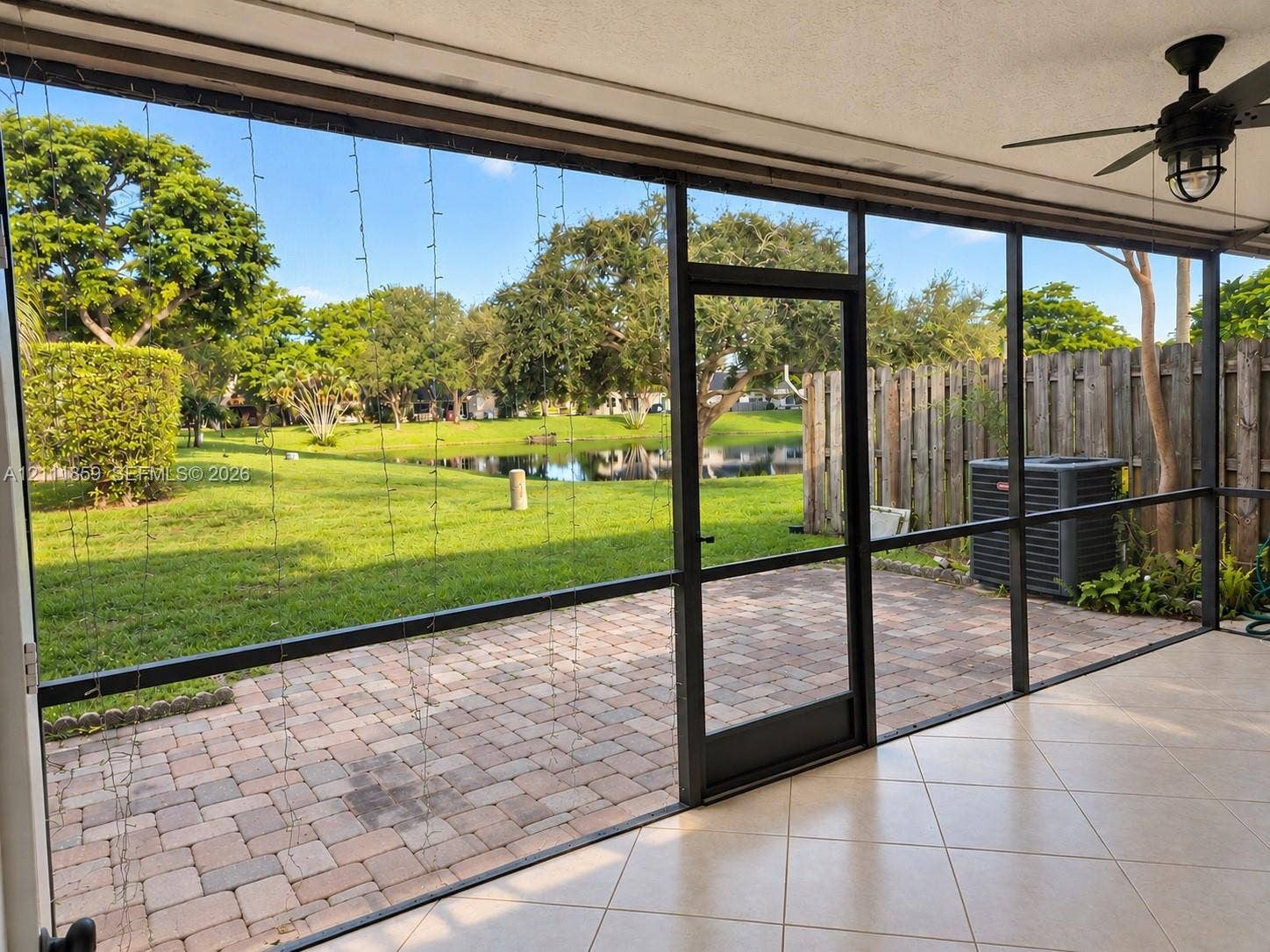 Screened-in patio with tiled flooring and ceiling-mounted fan, featuring brick-paver hardscaping, mature landscaping, and waterfront scenery