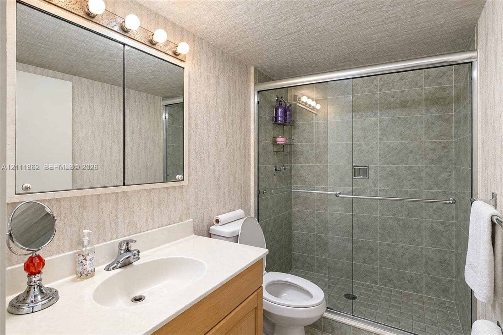 2030 S Ocean Drive, #2004, Hallandale Beach, FL 33009 Bathroom featuring a wood-finish vanity with a solid-surface countertop, an integrated sink, a mirrored medicine cabinet with integrated lighting, and a walk-in shower with a glass sliding door and tiled surround