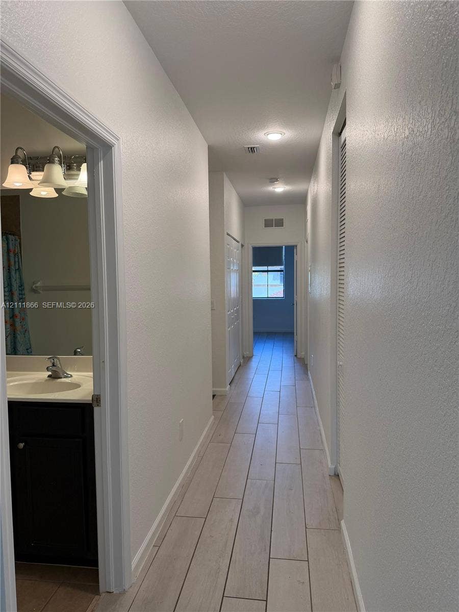 Hallway featuring wood-finish tile flooring, recessed lighting, and a bathroom containing a dark-toned vanity with a white countertop and multi-bulb light fixture