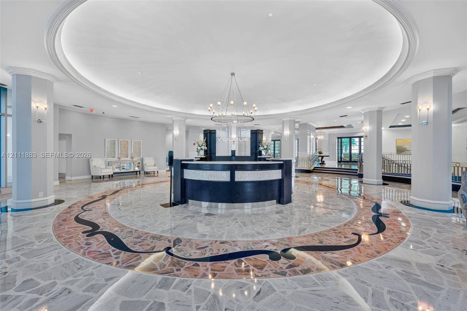 Circular lobby featuring an intricate stone-inlay floor, high coffered ceilings, structural pillars, a central built-in reception desk, and a tiered chandelier