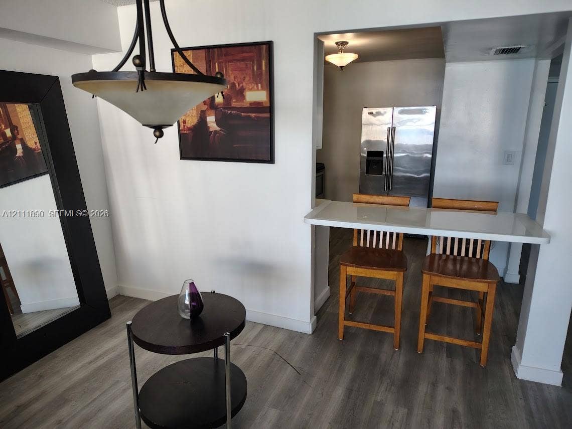 Dining area featuring a modern multi-arm pendant light and wood-finish flooring, complemented by a pass-through breakfast bar with a solid surface countertop and stainless steel refrigerator