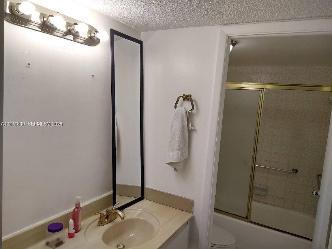 Bathroom featuring a vanity with integrated sink, brass-tone hardware, a vertical rectangular mirror, four-bulb vanity lighting fixture, and a tub-shower combination with a gold-tone sliding frame
