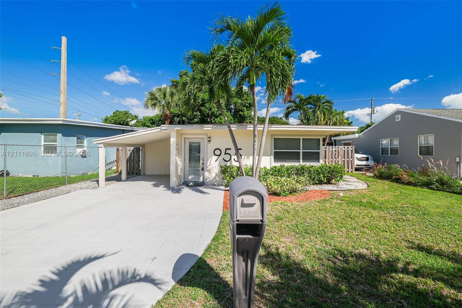 Single-story exterior featuring a concrete carport, front-facing windows with frame detailing, landscaped greenery, and mature palm trees