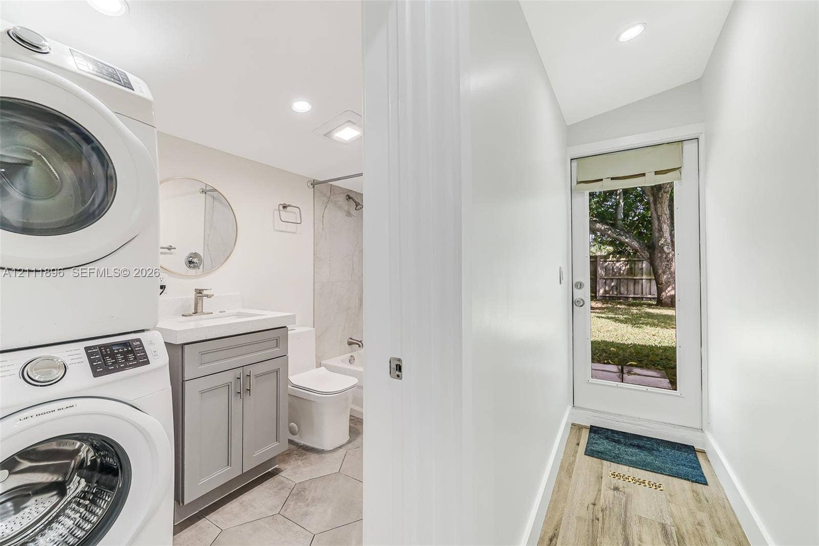 Combined utility and bathroom featuring a stacked washer and dryer, gray vanity with a rectangular basin, tile flooring, and exterior door with glass insert
