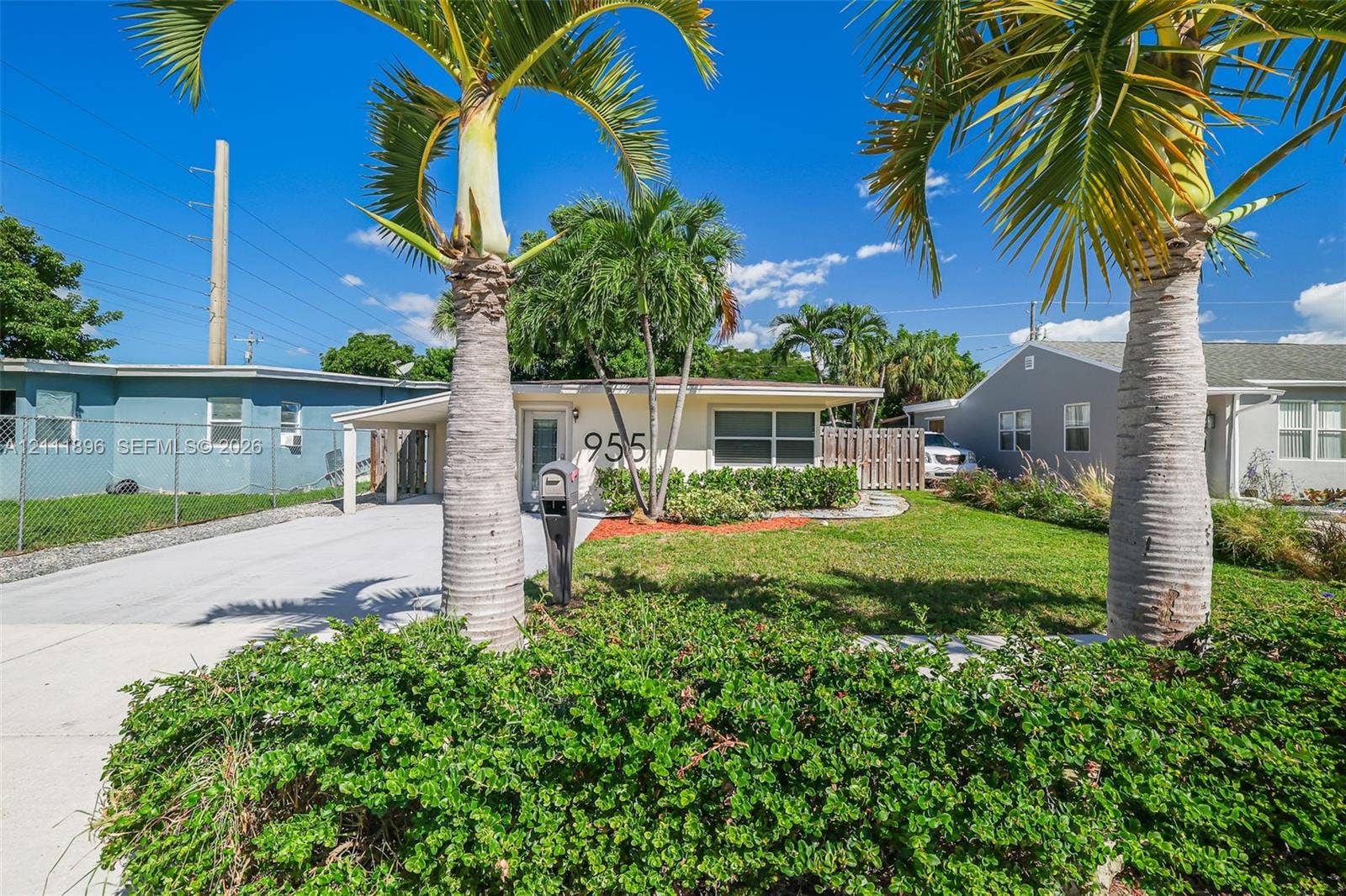 Single-story residence featuring a paved carport, mature palm trees, manicured landscaping, and a wood privacy fence