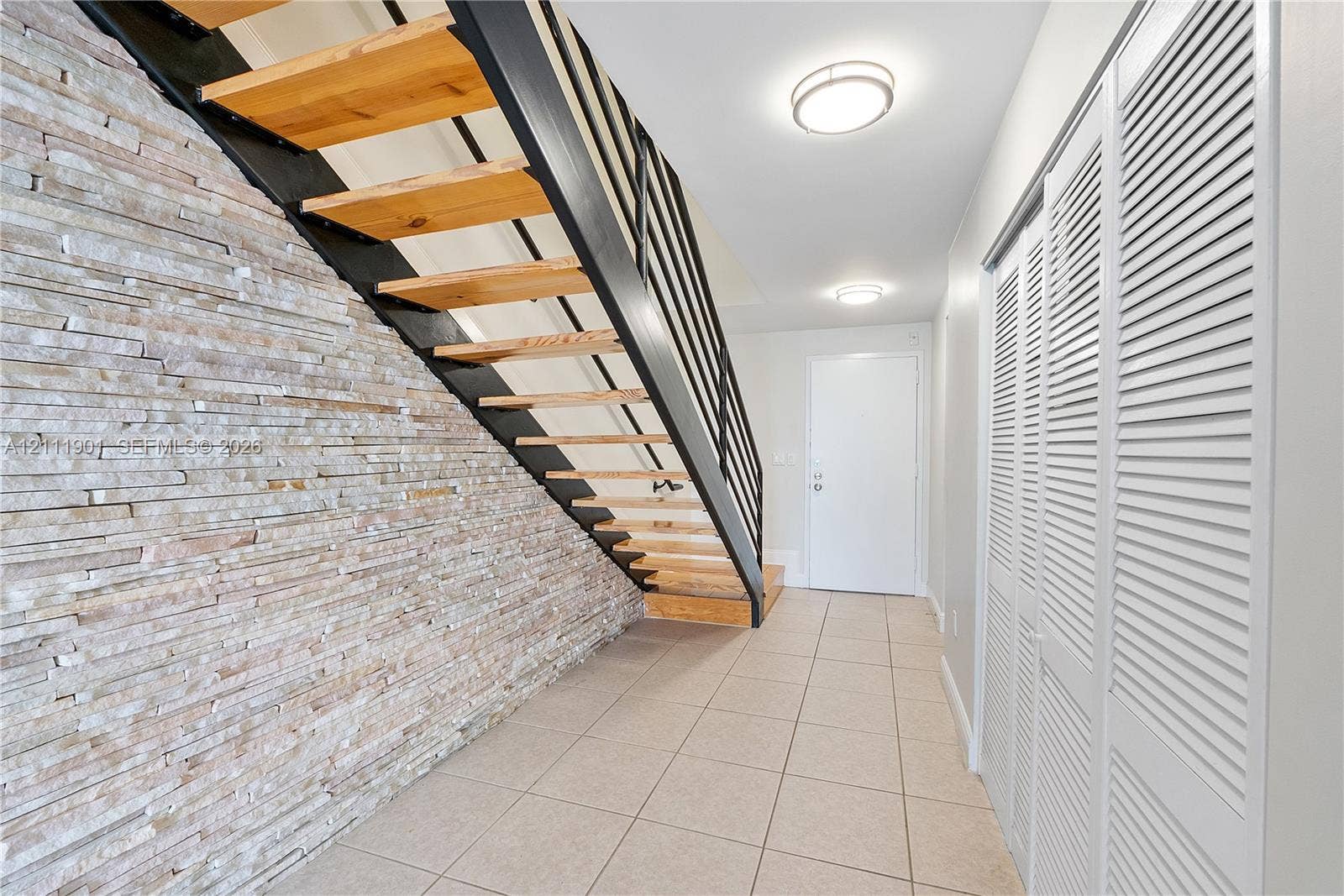 2977 Mcfarlane Road, #9, Miami, FL 33133 Open-riser staircase featuring natural wood treads, black metal railing, and a stacked stone accent wall, complemented by tile flooring and louvered double-door storage