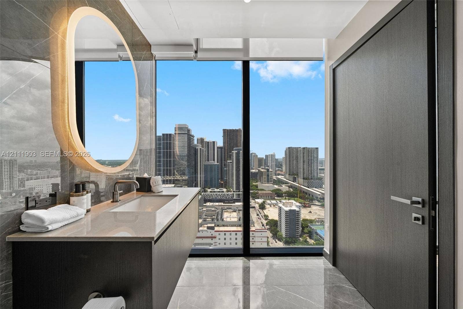 20 NE 11th Street, #4308, Miami, FL 33132 Floating vanity featuring a stone countertop, integrated sink, and wall-mounted faucet, complemented by an oval backlit mirror, full-height window, and polished stone flooring