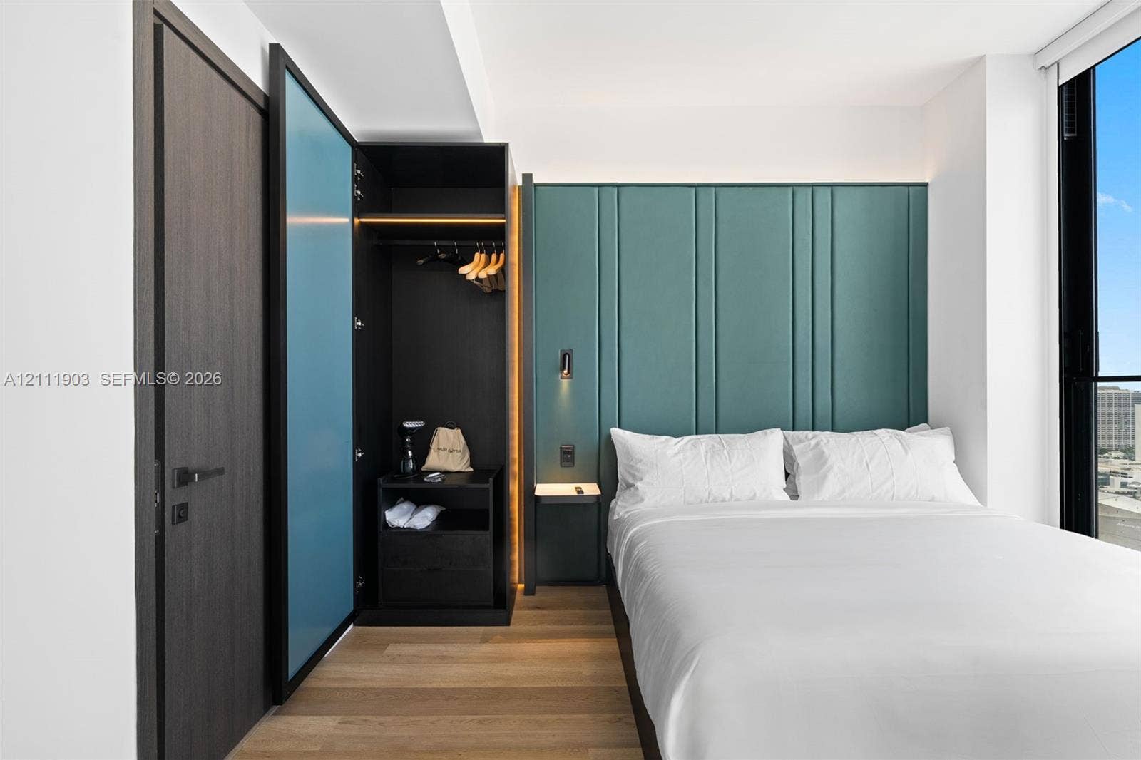 20 NE 11th Street, #4308, Miami, FL 33132 Full-height floor-to-ceiling windows, custom teal-paneled accent wall, wood-finish flooring, built-in wardrobe with integrated accent lighting, and modern matte black door hardware