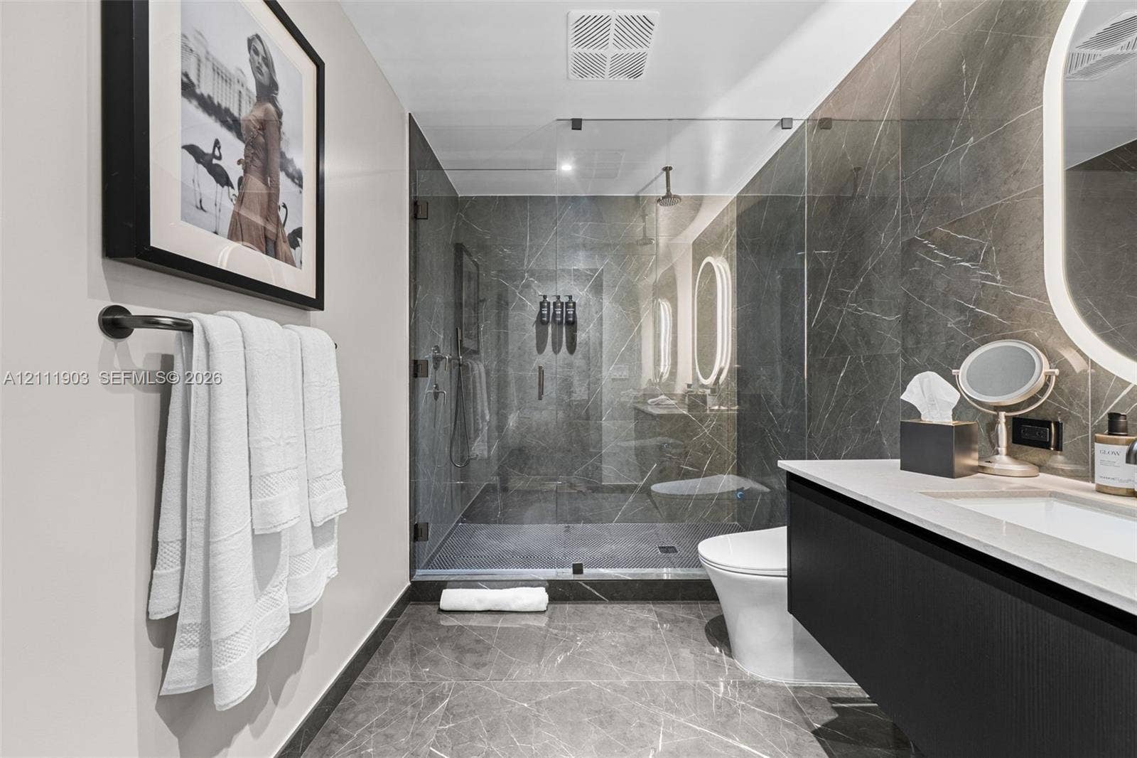20 NE 11th Street, #4308, Miami, FL 33132 Walk-in glass-enclosed shower featuring stone-finish wall and floor tile, wall-mounted matte black towel bar, floating vanity with solid-surface countertop, and oval LED-backlit mirror