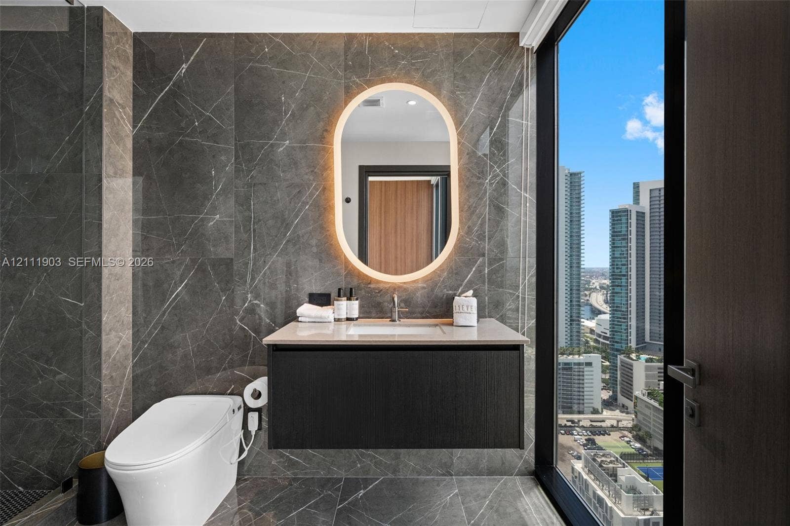20 NE 11th Street, #4308, Miami, FL 33132 Full-height stone wall tiling, modern wall-mounted vanity with a stone countertop, oval LED-backlit mirror, floor-to-ceiling windows, and wall-mounted toilet