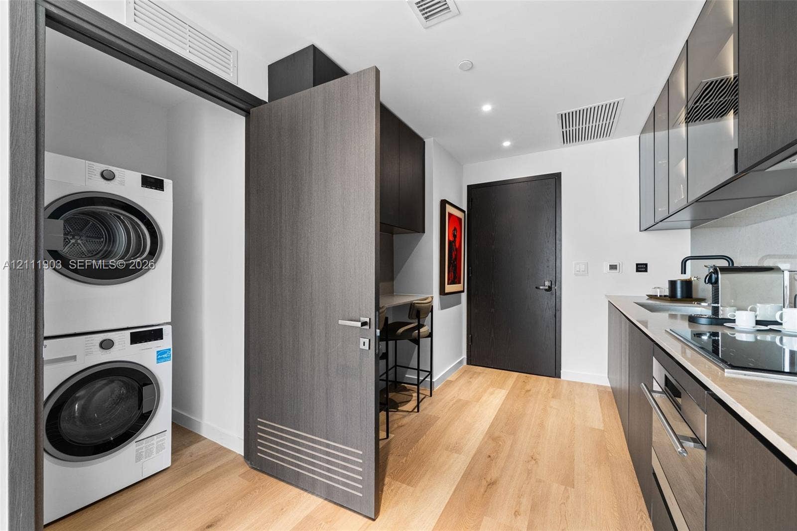 20 NE 11th Street, #4308, Miami, FL 33132 Contemporary kitchen featuring wood-finish flooring, dark-toned flat-panel cabinetry, light-toned countertops, an integrated cooktop, and an enclosed laundry area with stacked appliances