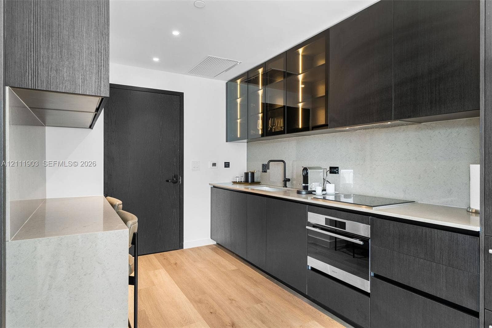 20 NE 11th Street, #4308, Miami, FL 33132 Modern kitchen featuring flat-panel dark cabinetry, stone-finish countertops, glass-front upper cabinets, a built-in stainless steel oven, and wood-finish flooring