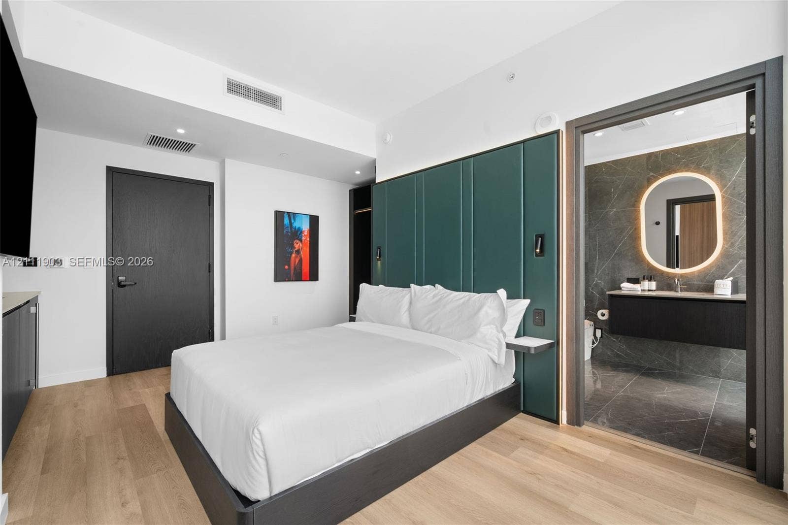 20 NE 11th Street, #4308, Miami, FL 33132 Custom floor-to-ceiling green panel headboard, wood-finish flooring, dark-toned interior doors, floating bathroom vanity, and rounded illuminated mirror