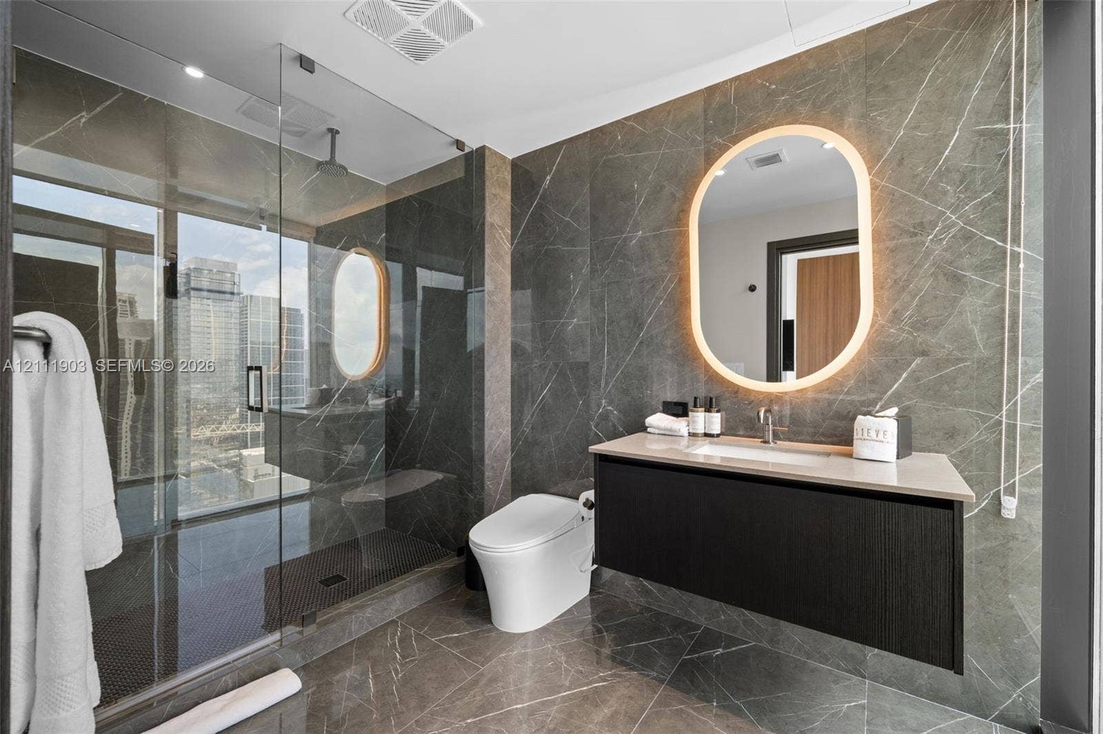 20 NE 11th Street, #4308, Miami, FL 33132 Full-height stone wall tiling, glass-enclosed walk-in shower, wall-mounted floating vanity, integrated stone countertop with undermount sink, and oval LED-backlit vanity mirror