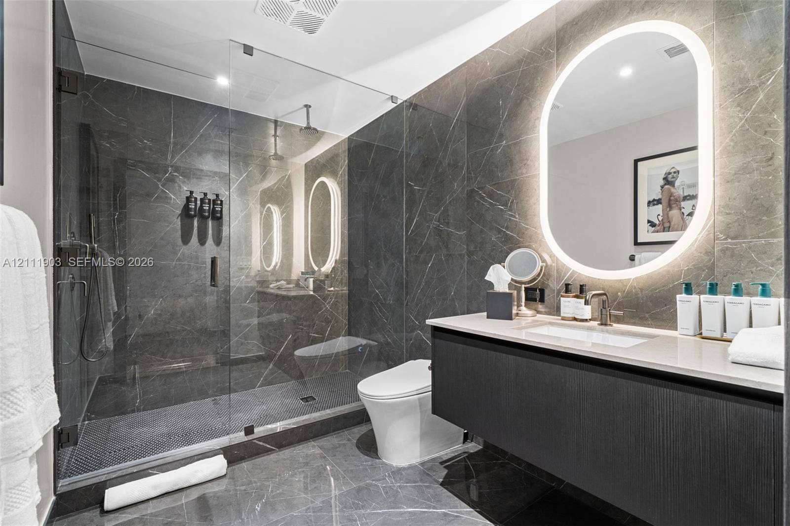 20 NE 11th Street, #4308, Miami, FL 33132 Full-height dark stone wall tiling, floating vanity with integrated countertop and sink, walk-in glass-enclosed shower with rainfall showerhead, and oval wall-mounted mirror with integrated backlighting
