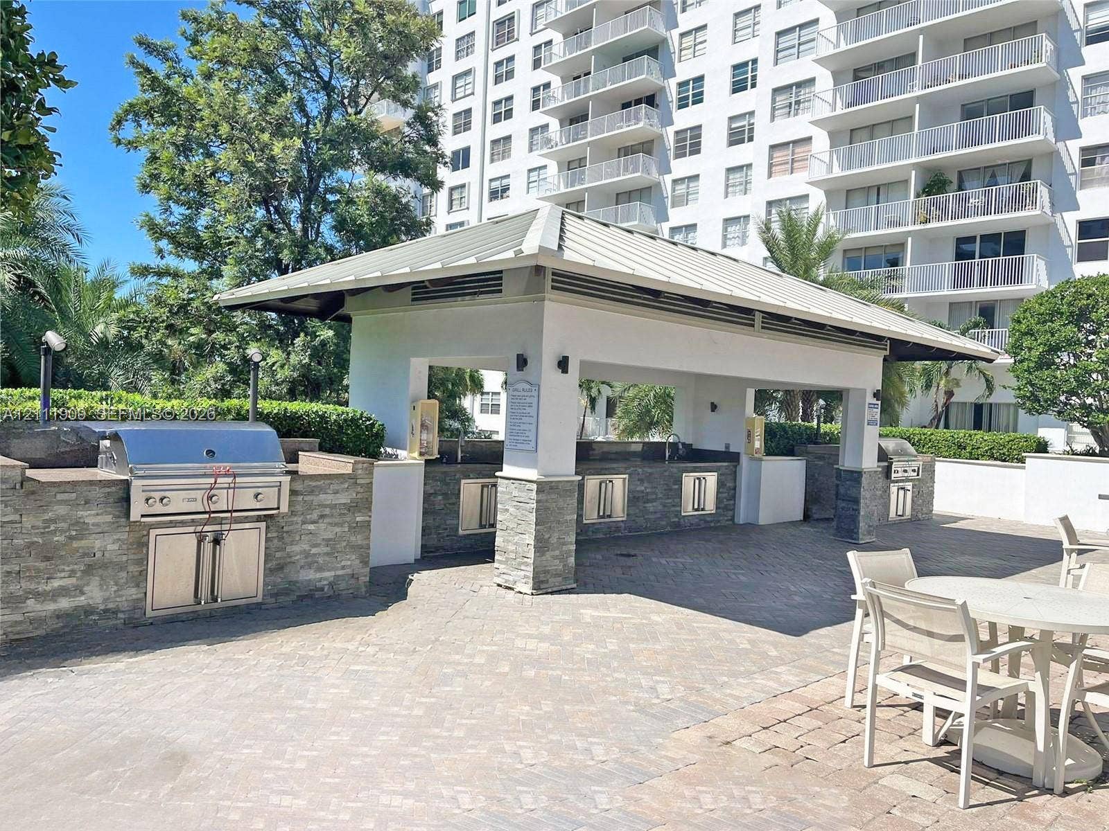 2851 NE 183rd Street, #1708E, Aventura, FL 33160 Covered outdoor kitchen featuring stone veneer base cabinetry, built-in stainless steel appliances, a metal roof structure, and brick paver flooring