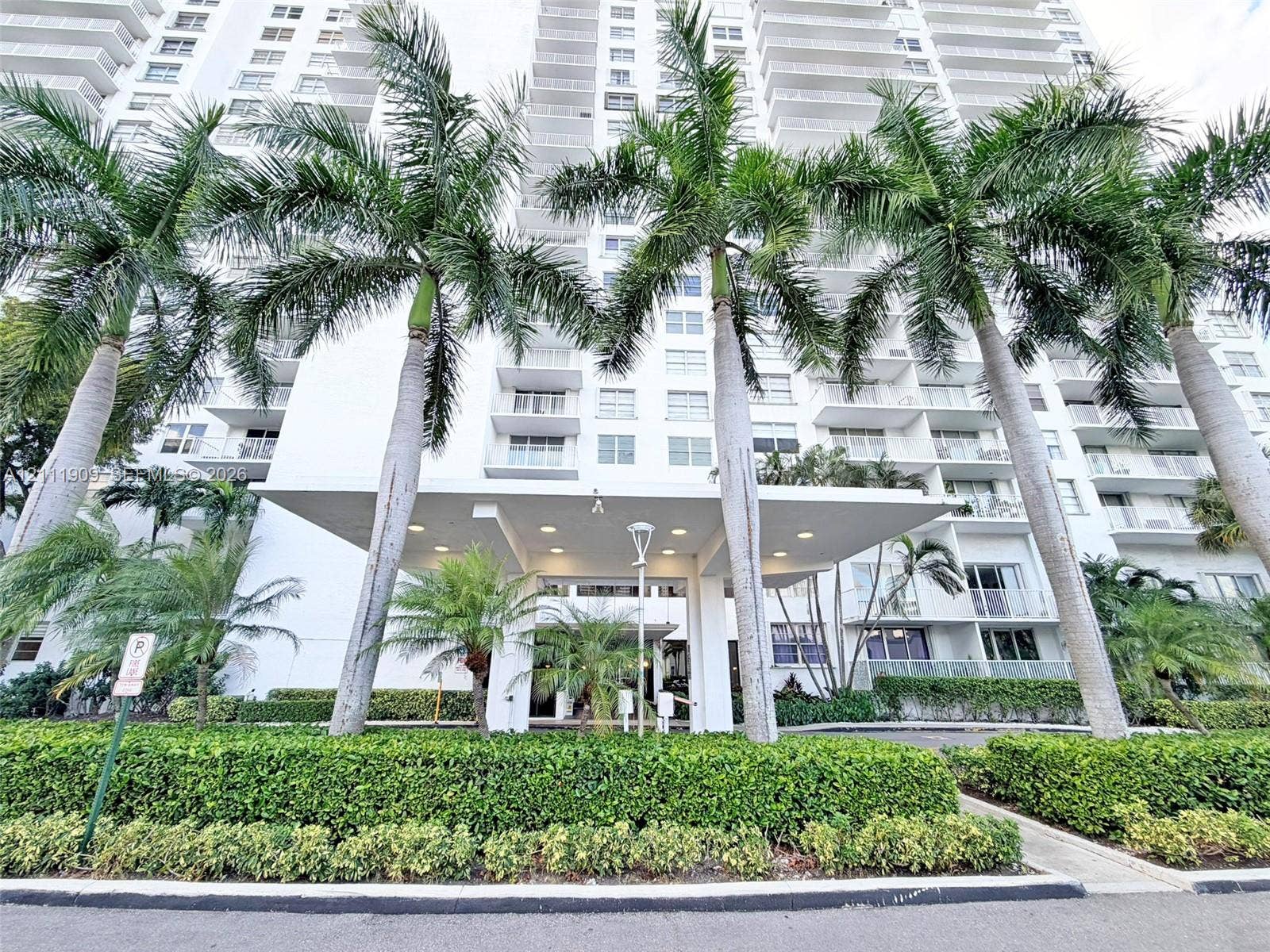 2851 NE 183rd Street, #1708E, Aventura, FL 33160 High-rise building exterior featuring a covered porte-cochere entry, wrap-around private balconies, recessed lighting, and manicured landscaping with mature palm trees