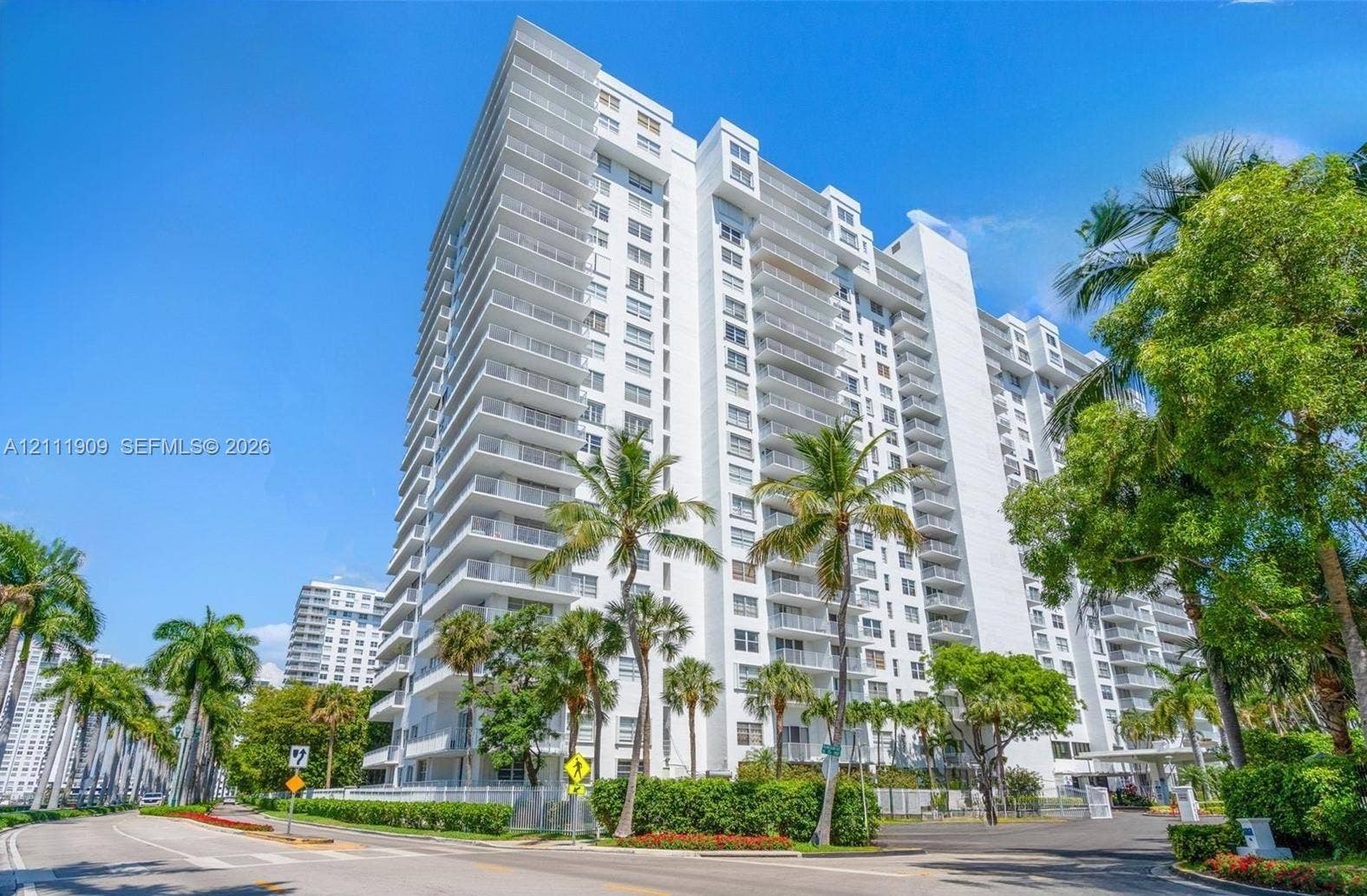 2851 NE 183rd Street, #1708E, Aventura, FL 33160 High-rise residential building featuring a white facade, wraparound balconies, glass railings, and manicured tropical landscaping