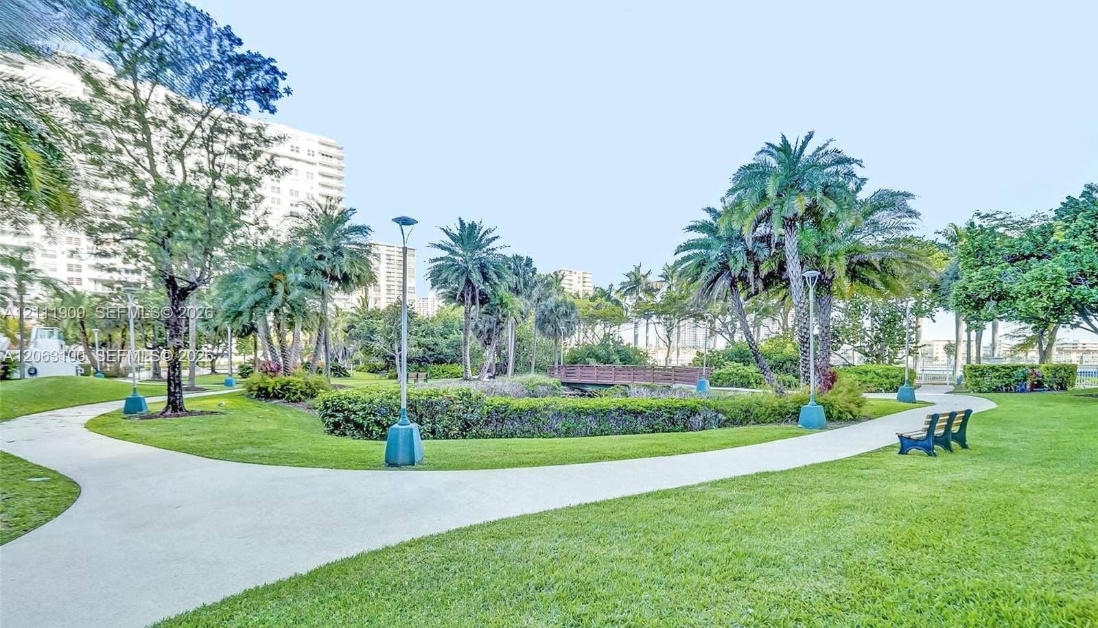 2851 NE 183rd Street, #1708E, Aventura, FL 33160 Landscaped grounds featuring winding concrete pathways, mature palm trees, decorative shrubbery, and a wooden pedestrian bridge