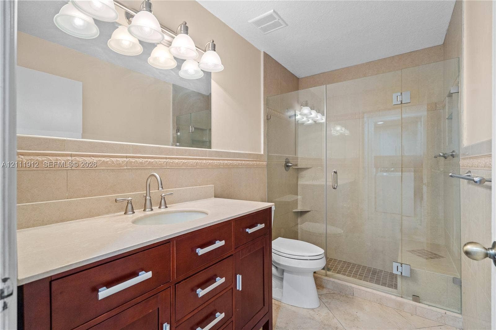 15681 SW 147th Court, Miami, FL 33187 Bathroom featuring a dark wood-finish vanity, light-toned countertop with an integrated sink, frameless glass-enclosed shower, decorative tile wall border, and multi-bulb vanity lighting