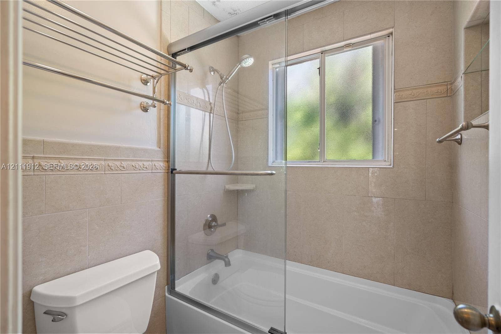 15681 SW 147th Court, Miami, FL 33187 Tub and shower combination featuring floor-to-ceiling tile surround with decorative inlay, glass sliding enclosure, handheld shower fixture, and wall-mounted metal shelving