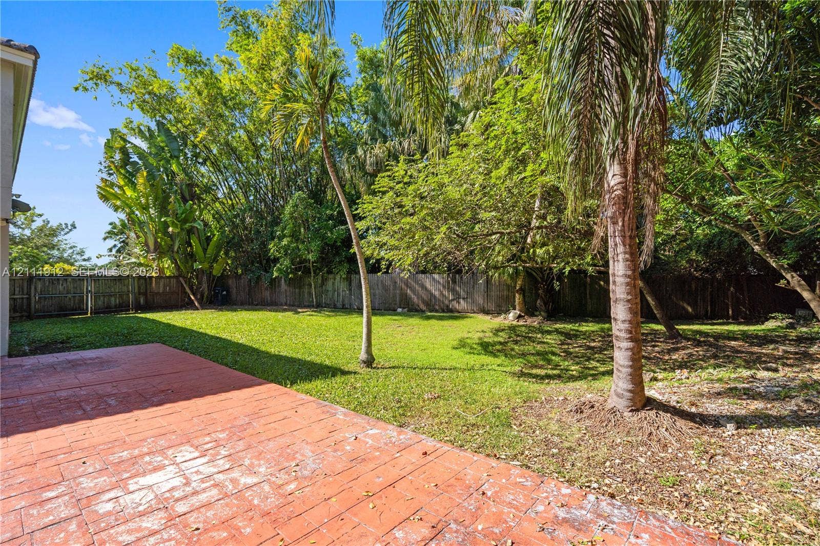 15681 SW 147th Court, Miami, FL 33187 Rear patio featuring stamped concrete, expansive lawn, perimeter wood fencing, and mature palms