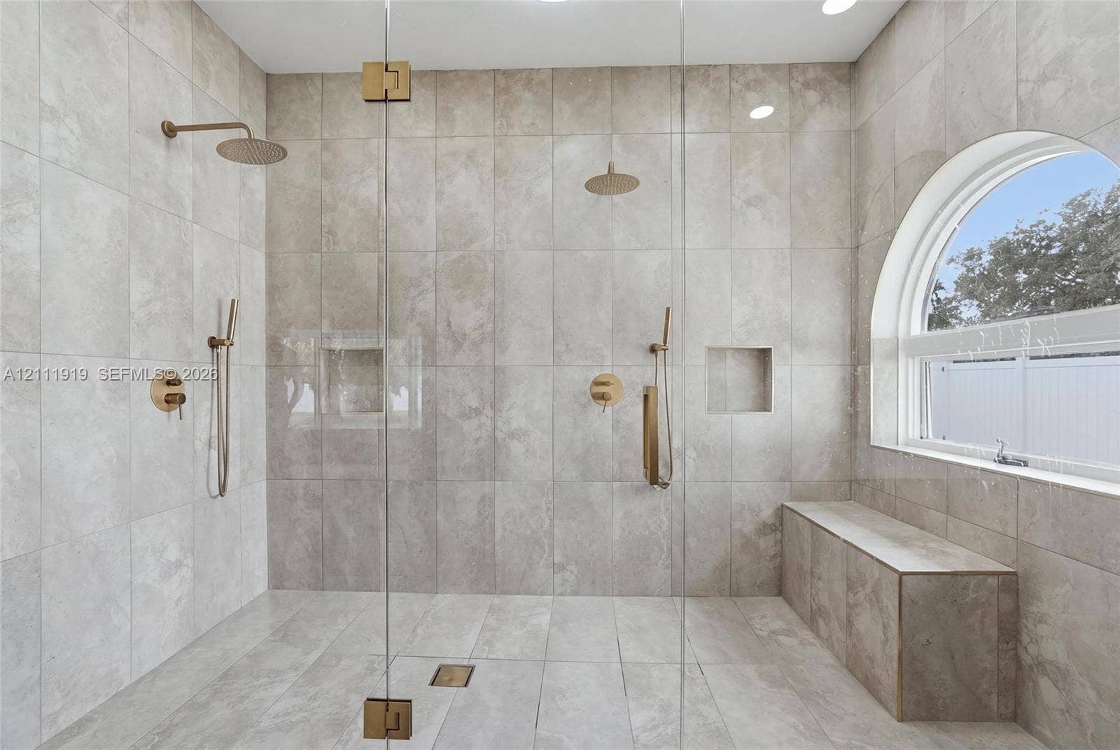 137 Arrowhead Lane, Haines City, FL 33844 Walk-in shower featuring floor-to-ceiling large-format neutral tile, dual rainfall showerheads with handheld sprayers, built-in recessed niches, a tiled bench, and a signature arched window