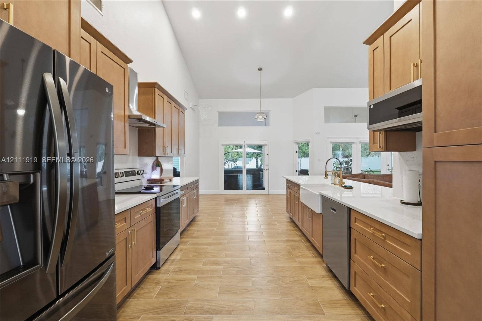 137 Arrowhead Lane, Haines City, FL 33844 Kitchen featuring light wood cabinetry with gold-tone hardware, white countertops, a farmhouse-style sink, stainless steel appliances, and wood-finish tile flooring under a vaulted ceiling