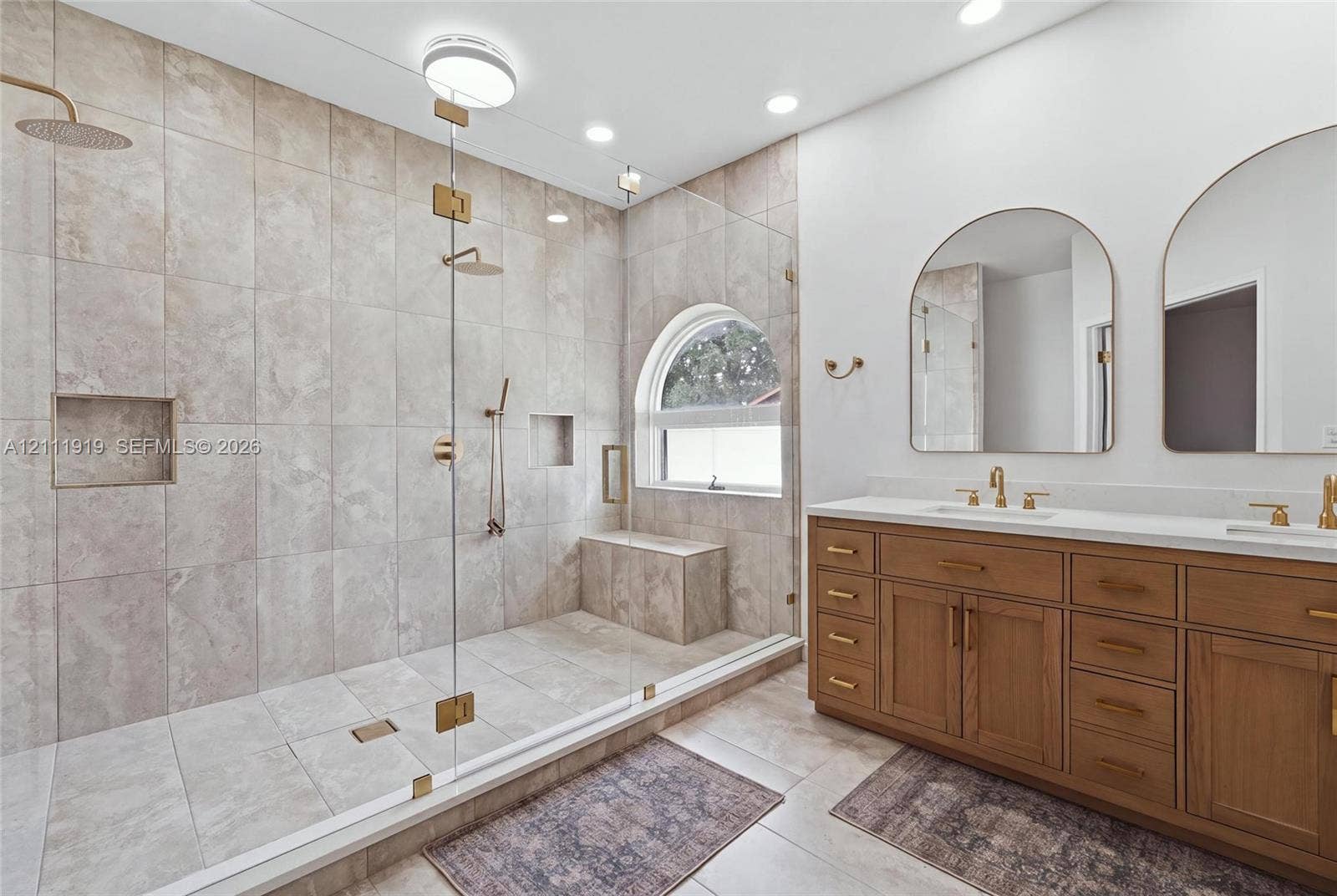137 Arrowhead Lane, Haines City, FL 33844 Walk-in shower featuring floor-to-ceiling stone-finish tile, an integrated bench, and a glass enclosure, paired with a dual-sink vanity with brass-finish hardware and arched mirrors