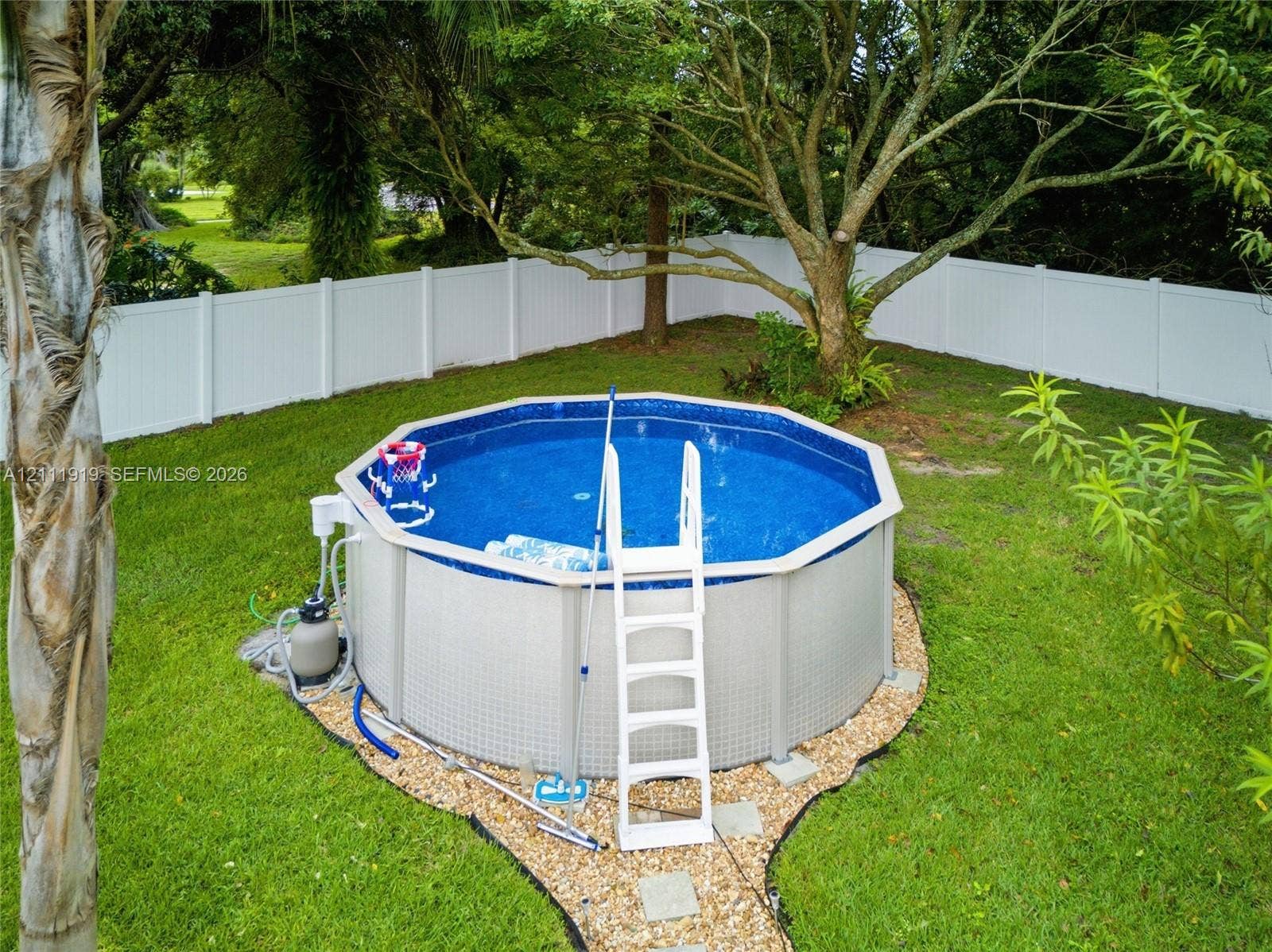 137 Arrowhead Lane, Haines City, FL 33844 Above-ground swimming pool with surrounding gravel border, enclosed by a white vinyl privacy fence and set within a grass-covered backyard featuring mature trees