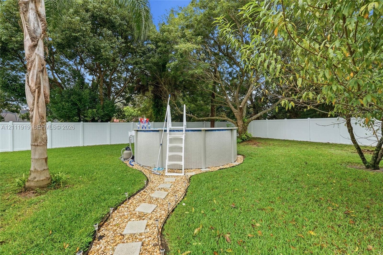 137 Arrowhead Lane, Haines City, FL 33844 Backyard featuring an above-ground swimming pool, white vinyl fencing, a gravel path with stone pavers, and mature trees