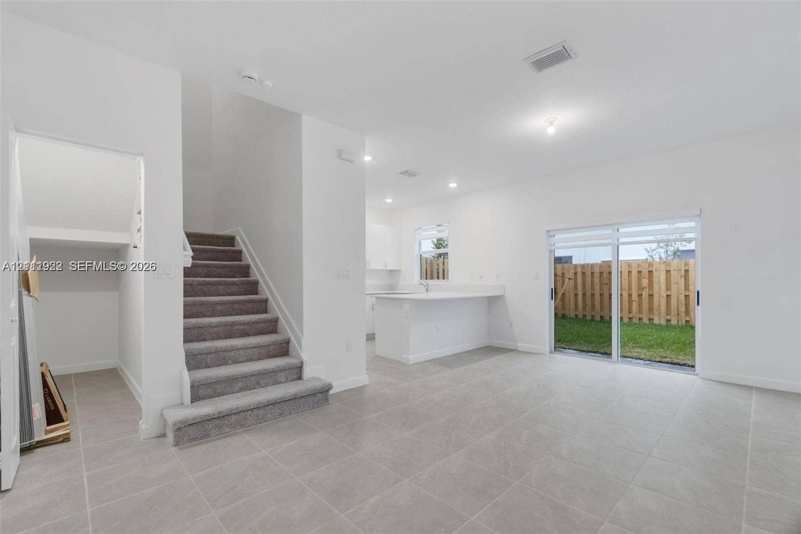 16281 SW 292nd Street, Homestead, FL 33033 Open-concept interior featuring large-format neutral tile flooring, carpeted staircase, kitchen with white cabinetry and breakfast bar, and sliding glass doors with access to a fenced rear yard