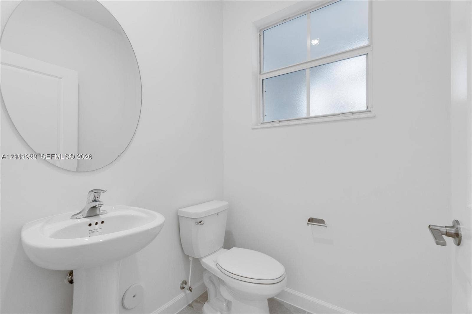 16281 SW 292nd Street, Homestead, FL 33033 Pedestal sink featuring a polished chrome faucet, round wall-mounted mirror, dual-flush toilet, and natural light via an obscured glass window
