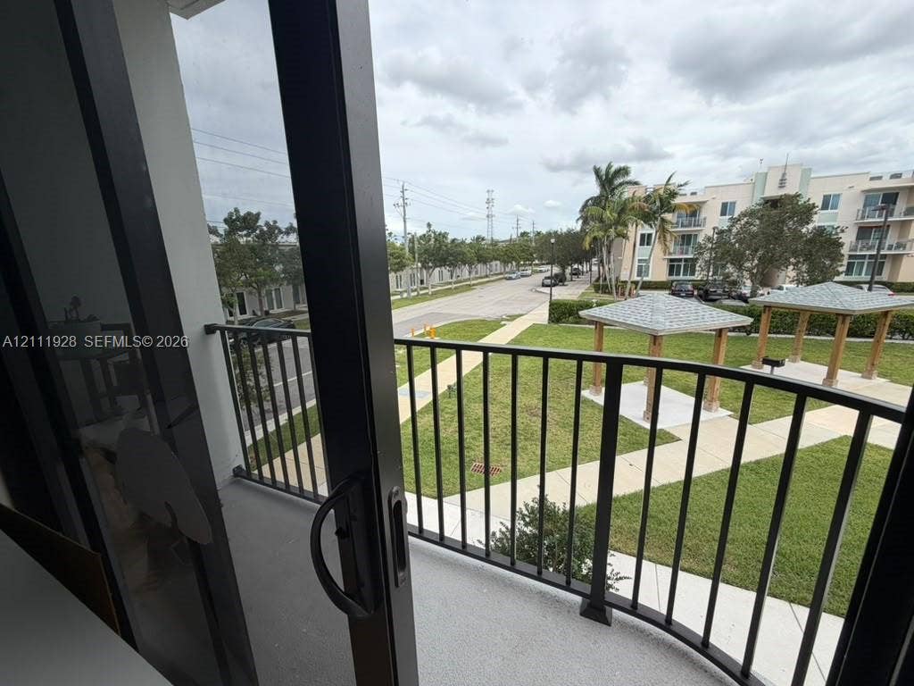 Balcony with black metal railing and concrete flooring, overlooking a green space featuring paved walkways and wooden-frame gazebos