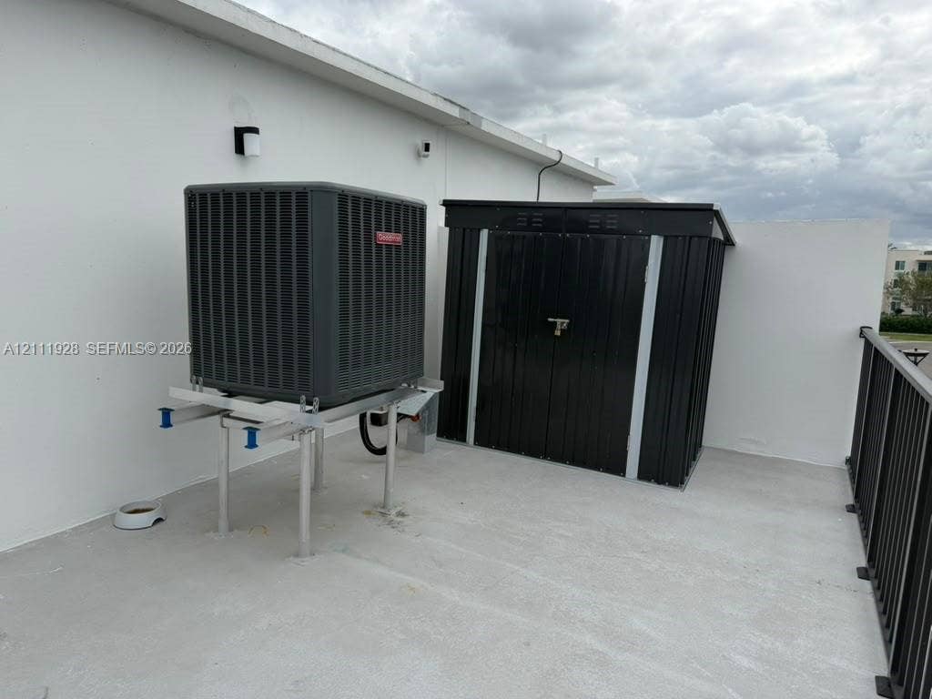 Exterior balcony featuring a concrete surface, metal privacy railing, wall-mounted exterior lighting, a stand-mounted HVAC condenser, and a secure metal storage unit