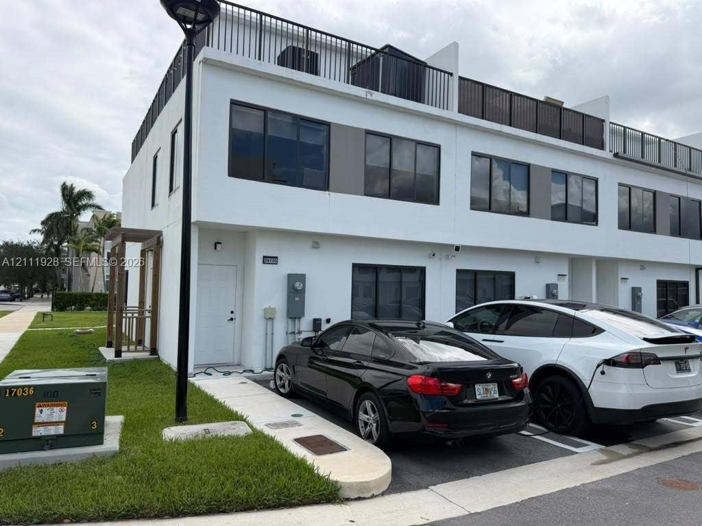 Modern two-story townhouse exterior featuring a smooth stucco facade, expansive rooftop terrace with black metal railing, large rectangular windows, and a paved private parking area