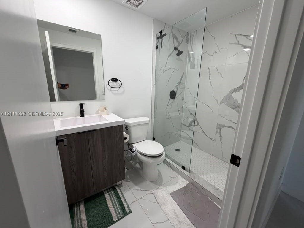 Modern bathroom featuring a wood-finish vanity, white countertop with integrated sink, walk-in shower with floor-to-ceiling veined tile, glass enclosure, and matte black fixtures