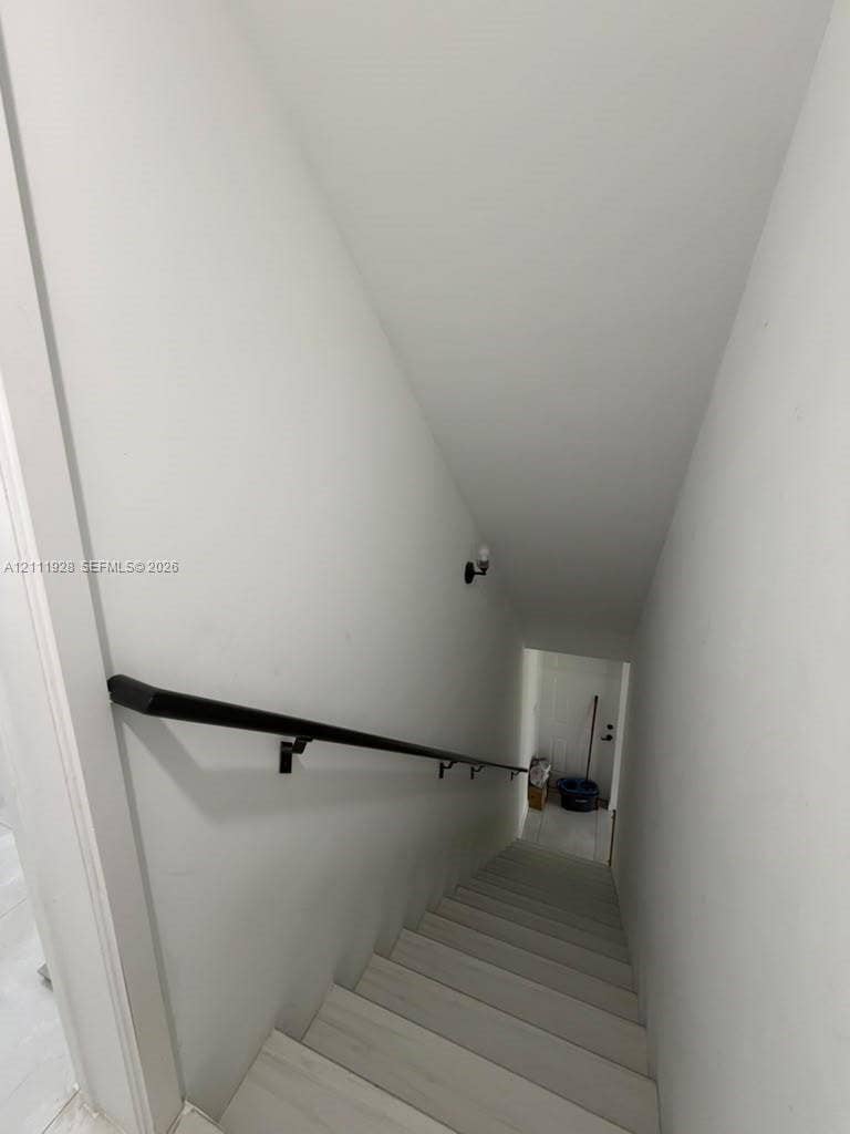 Staircase featuring light-toned wood-finish treads, a matte black wall-mounted handrail, and a singular wall-mounted sconce