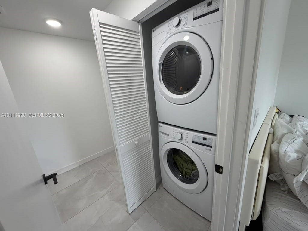 Stacked white front-load washer and dryer unit enclosed by white louvered bifold doors, featuring tile flooring and recessed ceiling lighting