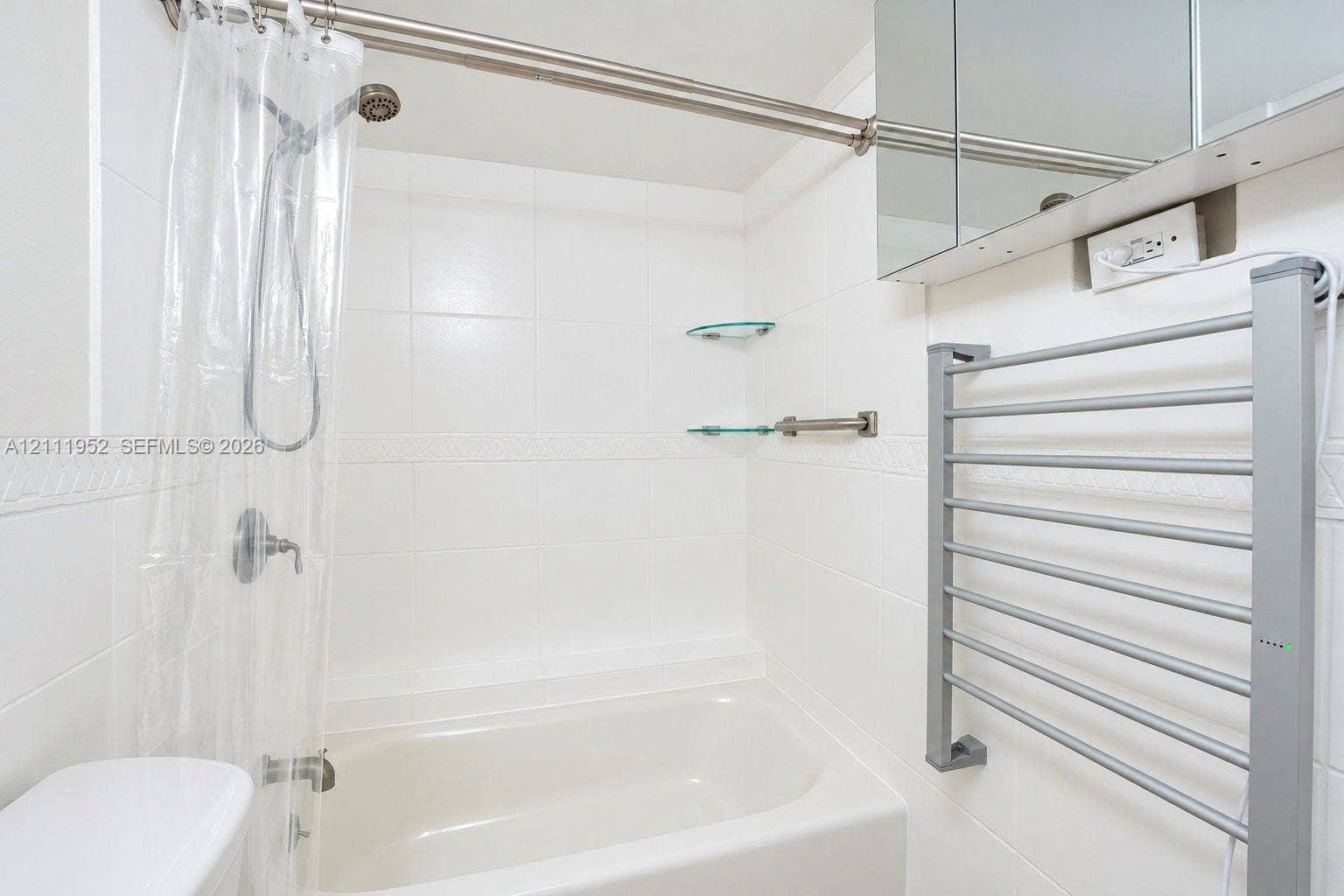 Full-size bathtub with white tile surround, integrated glass shelving, wall-mounted metal heated towel rack, and mirrored medicine cabinet