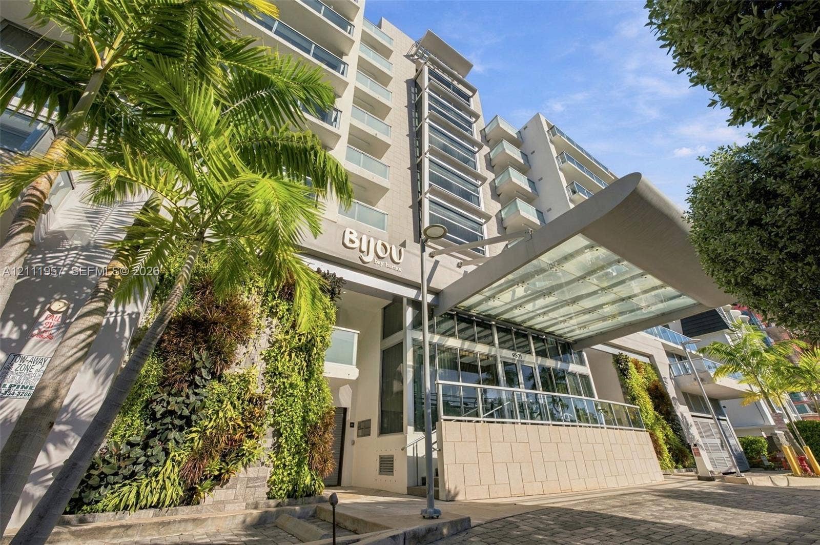 Multi-story residential building exterior featuring a glass-canopy entryway, tiered balconies with glass railings, vertical gardens, and stone-tiled facade elements