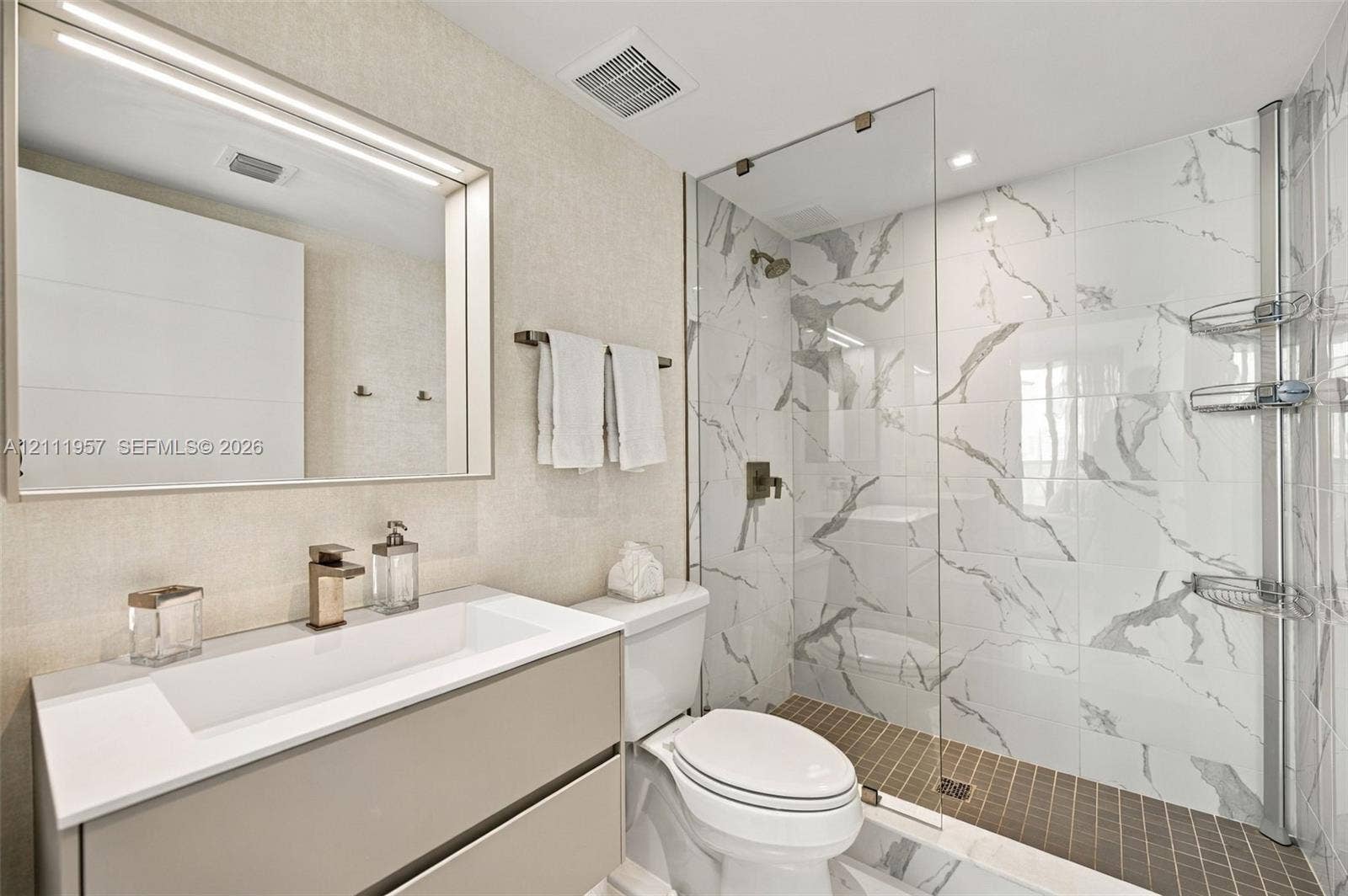 Floating vanity featuring a rectangular integrated sink, quartz countertop, and gold-tone faucet, complemented by a large framed vanity mirror, glass-enclosed walk-in shower with stone-patterned tile walls, and recessed lighting