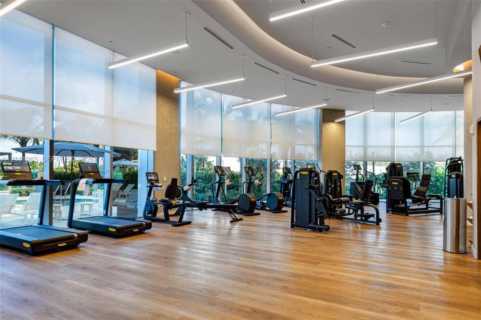 Fitness center featuring wood-finish flooring, floor-to-ceiling windows with roller shades, recessed cove lighting, suspended linear light fixtures, and a structural column