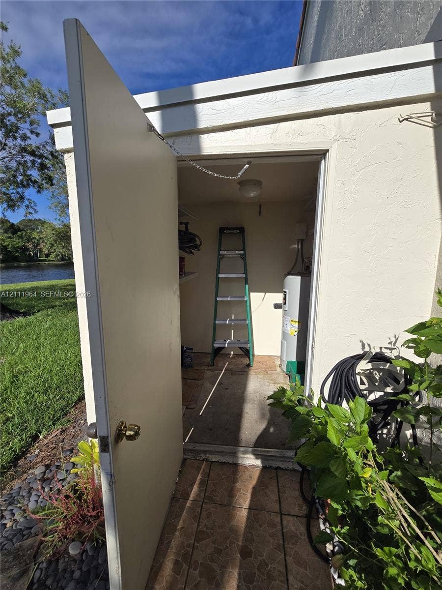 Exterior storage closet featuring smooth stucco walls, tiled flooring, a ceiling-mounted light fixture, and an attached water heater