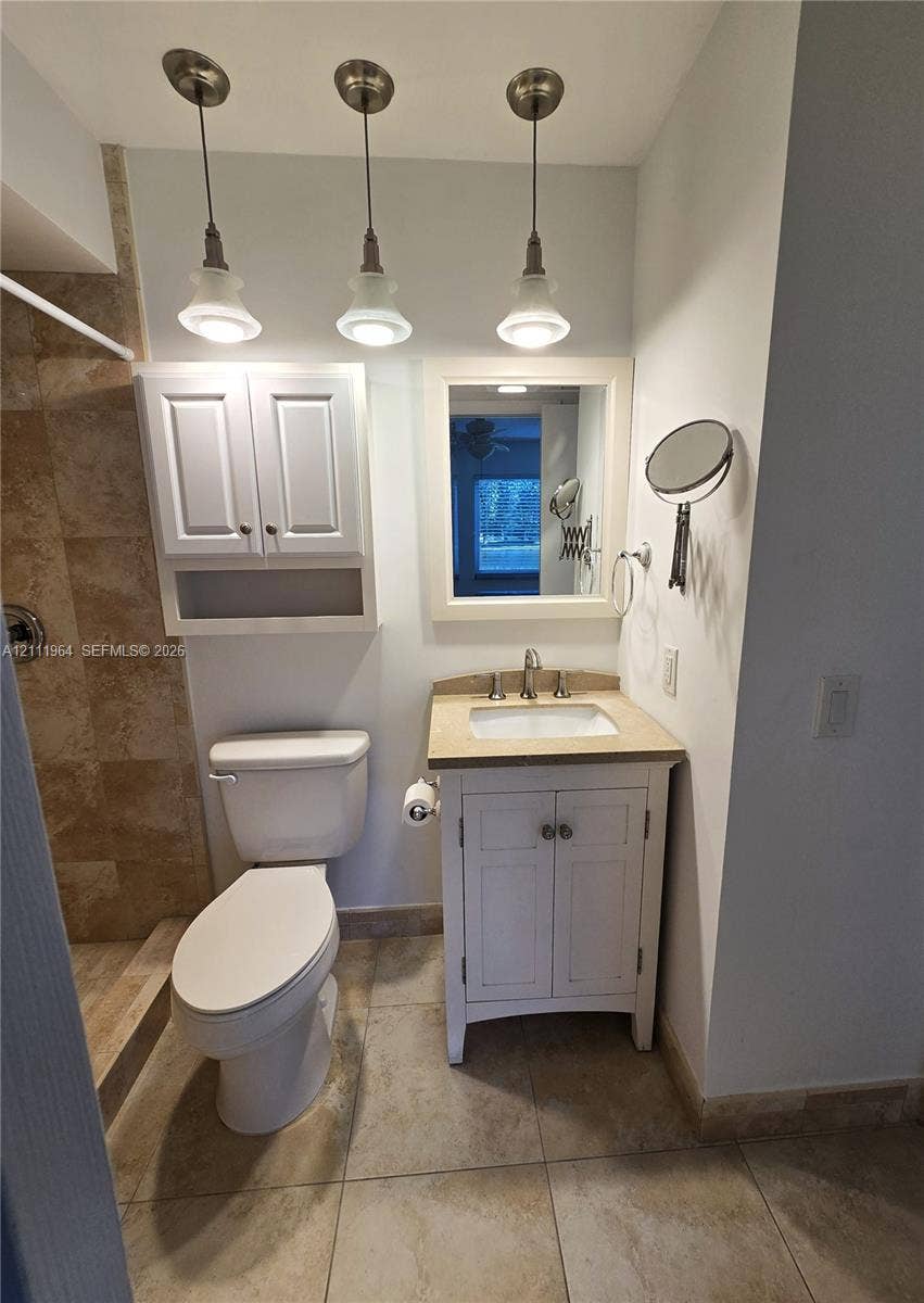 Bathroom featuring a vanity with an integrated sink and stone countertop, three brushed-nickel pendant lights, white cabinetry, a walk-in shower with tiled surround, and ceramic tile flooring