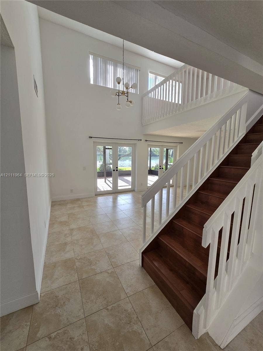 Two-story volume ceiling featuring a multi-light chandelier, staircase with wood-finish treads and white balusters, ceramic tile flooring, and dual sets of French doors