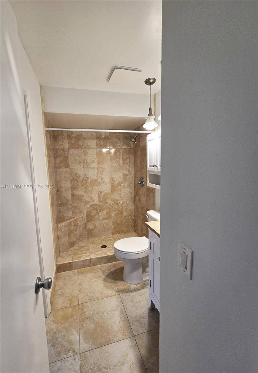 Walk-in shower with stone-tile surround, neutral ceramic tile flooring, white vanity cabinet, and pendant lighting